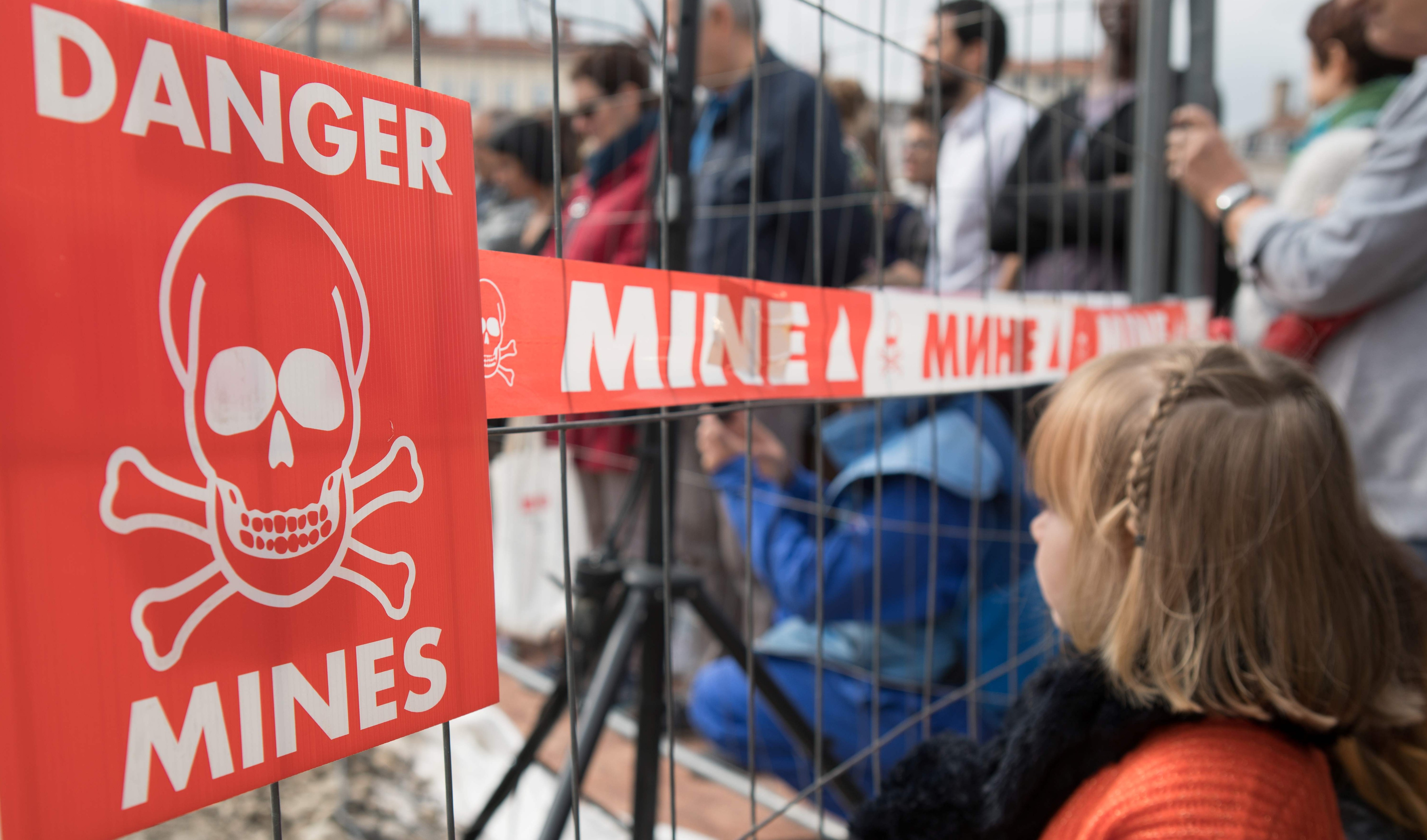 a little girl stands in front of a barrier fence. on the fence hangs a sign with a skull and crossbones and the words: DANGER MINES