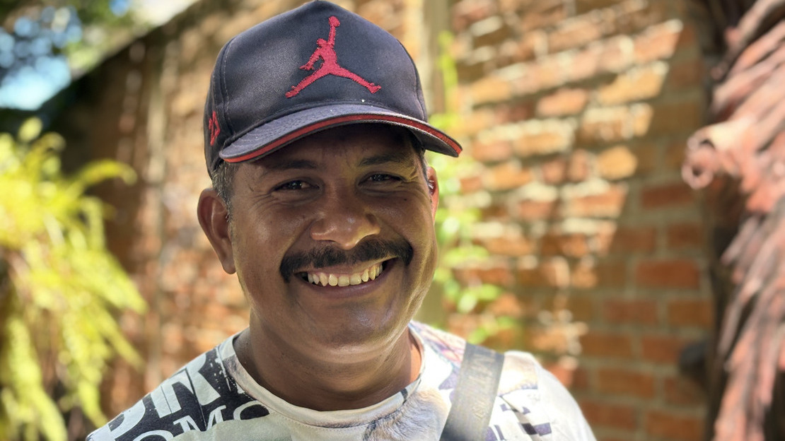 Close-up portrait of Julio César looking into the camera and smiling. He wears a cap and poses in front of a red brick wall. We can make out some vegetation behind him.