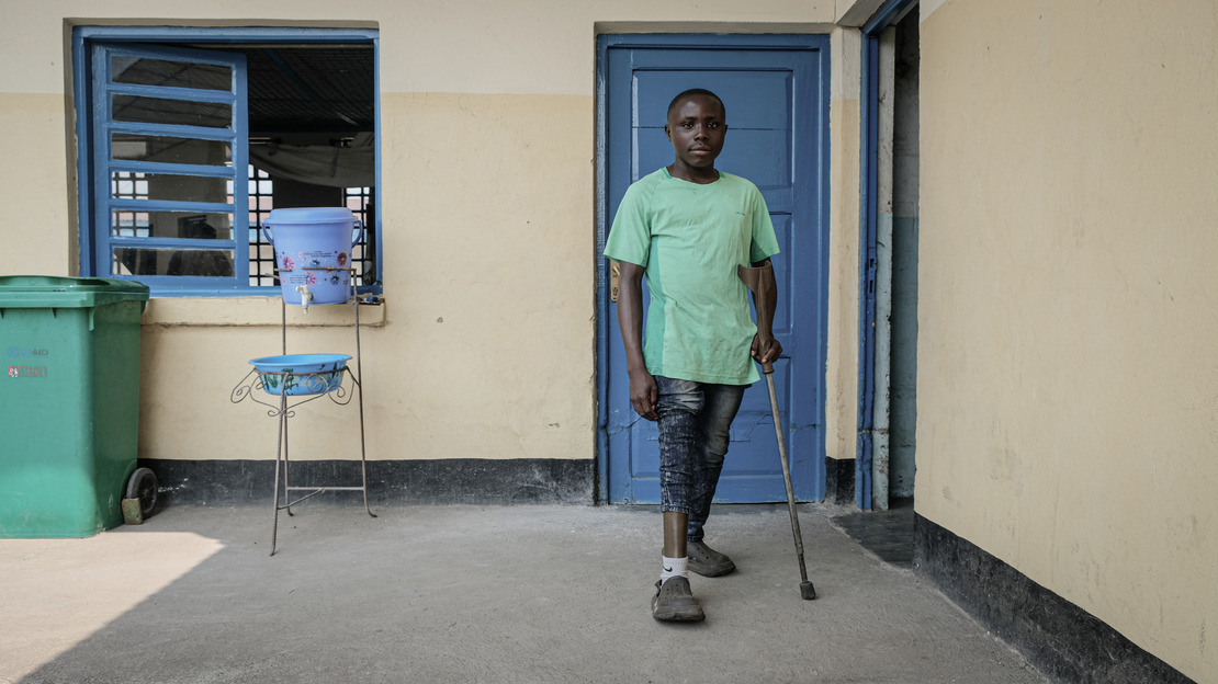 A man stands on a crutch. His right trouser leg is raised to reveal a prosthesis. Behind him, a wall with a window and two doors.