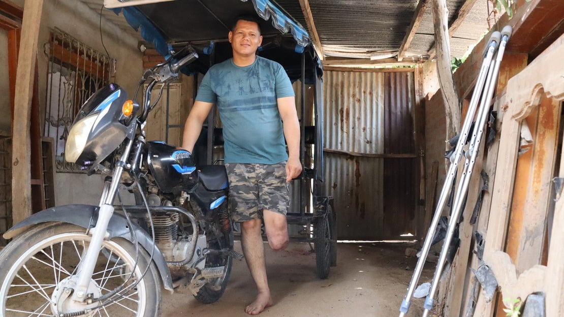 In a wooden and tin shed, a man stands, leaning on a motorcycle connected to a cariole with a seat. The man smiles, his left leg cut off below the knee.