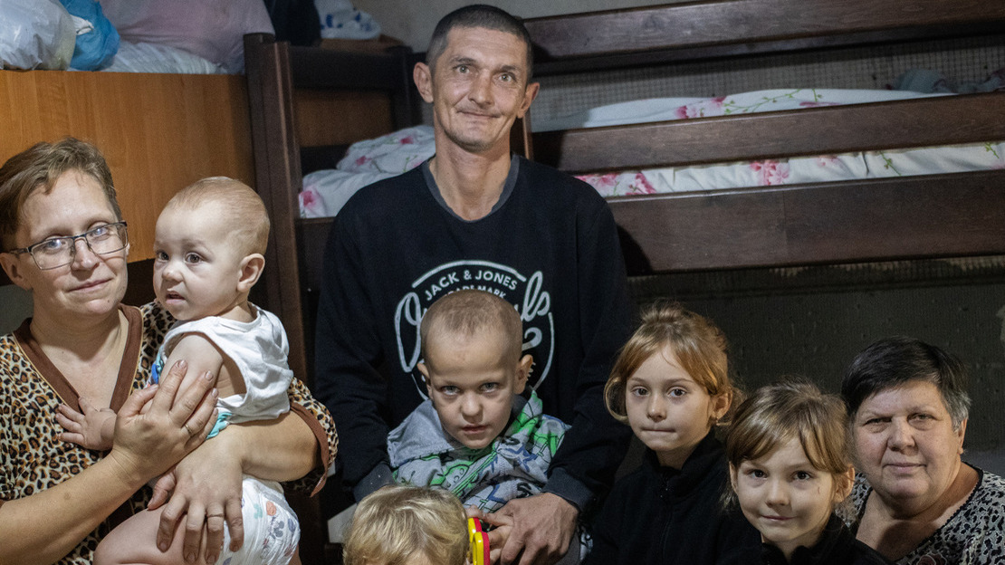 A large family is gathered in a room with bunk beds in the background. The father, in the center, has a gentle smile, while the mother, on the left, holds a baby in her arms. Around them, several children and their grandmother sit or stand, looking at the camera with shy smiles. 