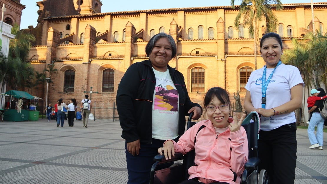 Trois femmes se tiennent sur une place, devant une cathédrale illuminée par le soleil. Deux d'entre elles sont debout et entourent la troisième, assises sur un fauteuil roulant. Elles sourient à la caméra.