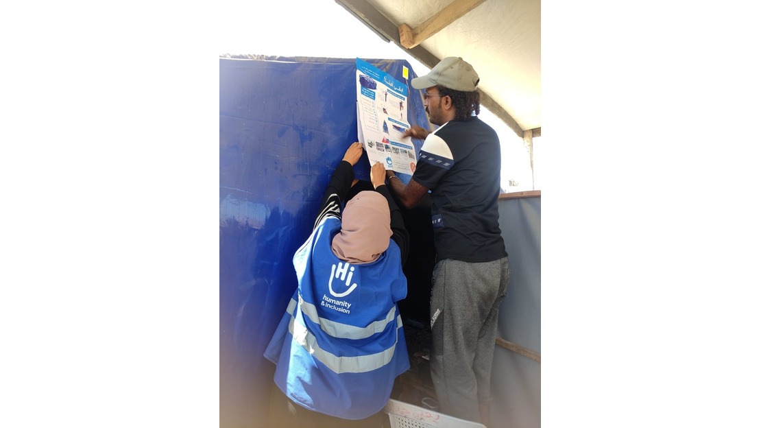 A HI staff woman, wearing a blue vest with the HI logo, and a man, are putting up a sign informing people about explosive remnants. They are in a refugee camp, sticking the sign on a wall. 