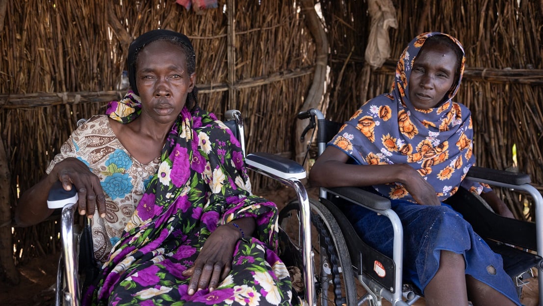 Kaltouma et Hassaneih sont côte à côte, chacun dans son fauteuil roulant. Elles sont abrités dans une maison en bois, et regardent l'objectif. Katlouma, à gauche, porte une robe avec un tissu à fleurs violet et blanc, tandis qu'Hassaneih, à droite, porte un foulard bleu à fleurs oranges. 