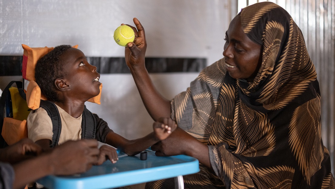 The photo is indoors, in an HI rehabilitation room. Omran, on the left, is in an adapted wheelchair and looks at his mother, who is showing him a tennis ball. Djimilla, his mother, looks at Omran with a smile. 