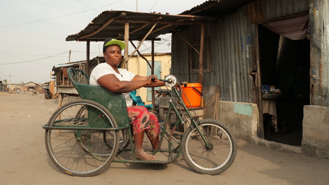 A woman sits on a tricycle, looking at the camera. She is in front of a shelter made of sheet metal and cement, and behind her we can make out a street and other buildings.