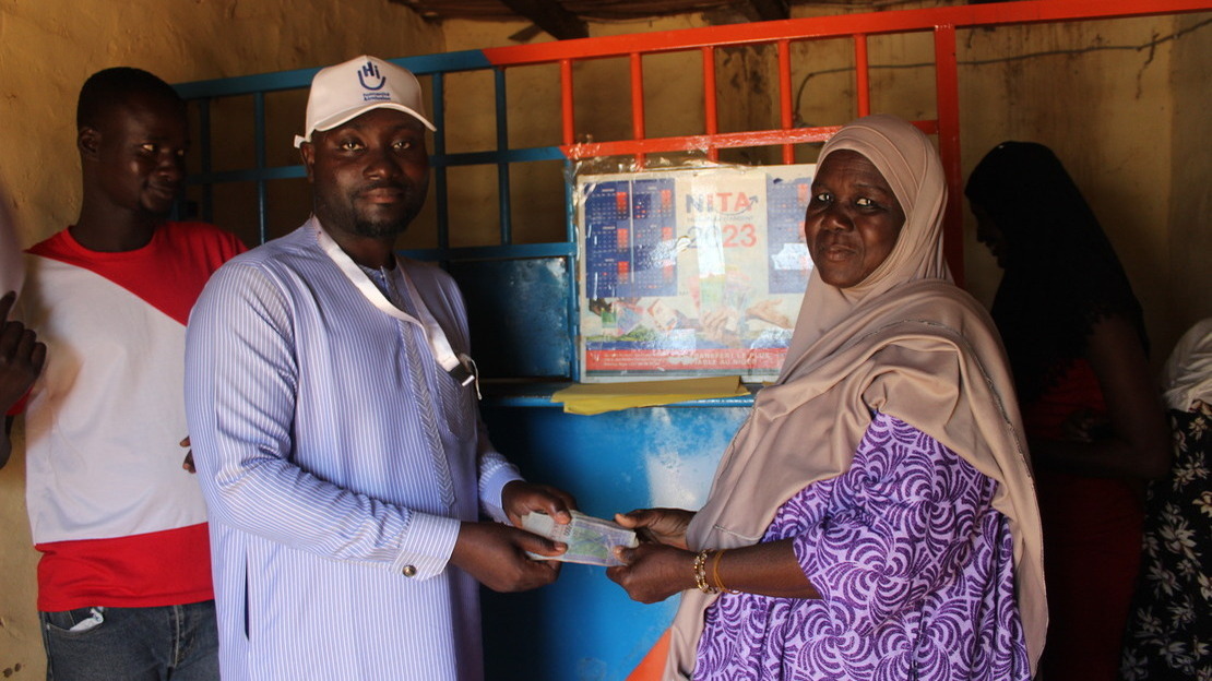 In front of a metal counter, two people face each other, faces turned towards the camera. On the left, a man wearing an HI cap, on the right, a woman. They are holding an envelope in all four hands.