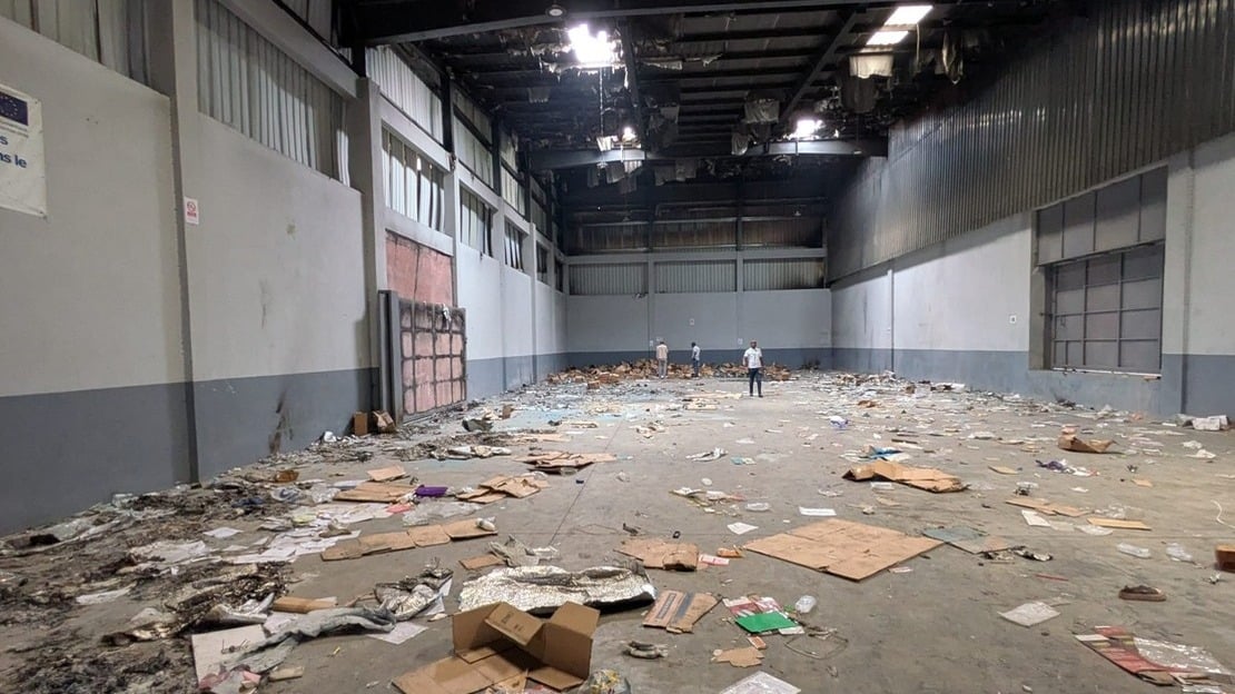 Inside a warehouse: the floor is littered with cardboard boxes, sacks and garbage of all kinds. Part of the roof is black with soot, and sheet metal panels are peeling off.