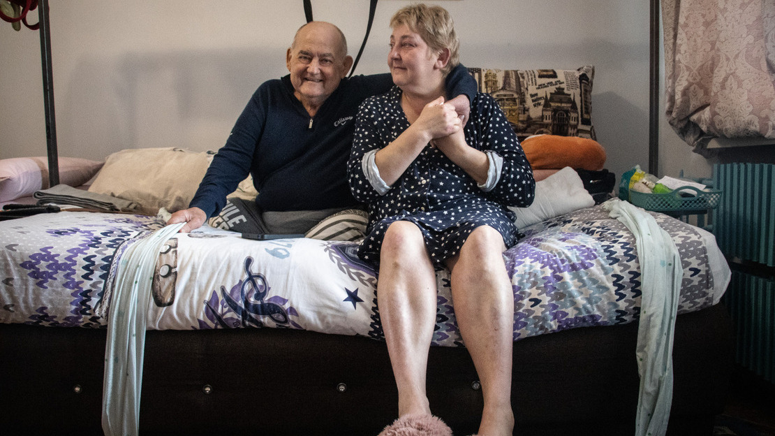 An elderly couple sits on a bed, sharing a moment of closeness. The man, dressed in a dark sweater, smiles warmly, while the woman, wearing a star-patterned robe and pink slippers, gently holds his hand over his shoulder. The intimate atmosphere of the room, with its colorful bedding and everyday objects, creates a feeling of warmth and connection.